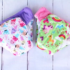 Może przedstawiać: Dwa wielorazowe pieluchy materiałowe z kolorowymi nadrukami. Jedna pielucha ma fioletowy pasek i białe tło z wzorami w kształcie lodów i chmur. Druga pielucha ma różowy pasek i zielone tło z wzorami w kształcie tęczy i jednorożców.