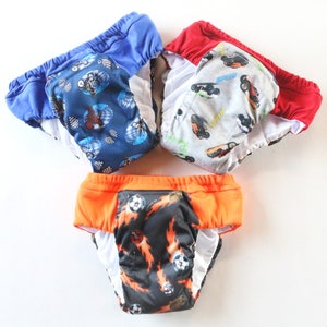 May include: Three colorful training pants with elastic waistbands. One is blue with a motorcycle pattern, another is red with a car pattern, and the third is orange with a soccer ball and flame design. The interior is white.