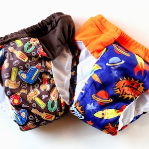 May include: Two reusable cloth training pants with colorful prints. One has a brown background with tools and the other has a blue background with rockets and planets. Both have orange elastic waistbands.