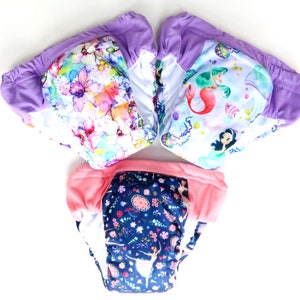 May include: Three cloth diapers with colorful patterns. One features a floral design, another a mermaid theme, and the third a ballerina print. Each diaper has a purple or pink trim and a white interior.
