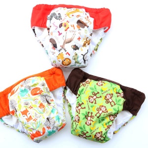 May include: Three reusable cloth training pants with different colorful animal prints. The first pant has a red waistband and a white background with a variety of animals and letters. The second pant has an orange waistband and a white background with a fox and floral print. The third pant has a brown waistband and a green background with monkeys and bananas.