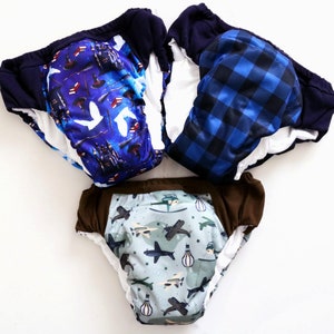 May include: Three reusable cloth training pants with different patterns. One has a blue and white checkered pattern, one has a blue and white pattern with a castle and owls, and one has a blue and green pattern with airplanes and hot air balloons.