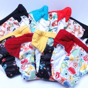 May include: A collection of eight reusable cloth diapers in various colors and patterns, including black with stars, yellow with planets, red with dinosaurs, and white with colorful creatures.