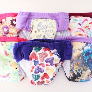 May include: A set of six colorful reusable training pants with various patterns, including donuts, unicorns, hearts, and galaxy designs. The pants are made of soft, breathable fabric and are designed to help toddlers transition from diapers to potty training.