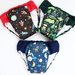 May include: Three reusable training pants with different colorful prints. One has a black background with rockets and stars, another has a green background with dinosaurs and plants, and the last one has a blue background with animals and stars.