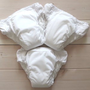 May include: Three white cloth diapers laid out flat on a wooden surface. The diapers are folded in half and have elastic around the legs and waist.