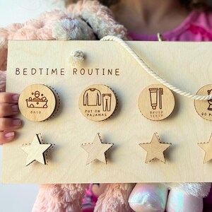 May include: Wooden bedtime routine chart with five circular icons for bath, pajamas, brushing teeth, potty, and story time. Includes five star-shaped reward tokens. The chart is held by a person.