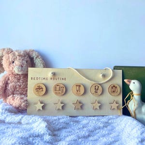 May include: A wooden bedtime routine chart with a rope handle. The chart has five circular icons with bedtime tasks and five star-shaped cutouts. A plush bunny and a decorative duck are in the background.