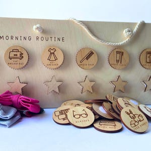 Pode incluir: Quadro de rotina matinal em madeira com as palavras "MORNING ROUTINE" no topo. Inclui círculos de madeira com ícones gravados para tarefas como comer o pequeno-almoço, vestir-se, escovar os dentes e muito mais. Tokens de madeira adicionais estão incluídos.
