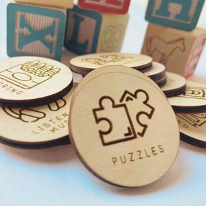 May include: A collection of circular wooden tokens featuring laser-engraved icons and text, including labels such as Coloring, Listen Music, and Puzzles. The tokens are arranged in a pile, with colorful wooden alphabet blocks visible in the background.