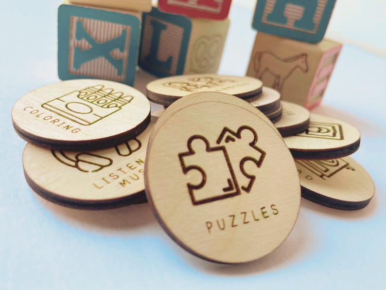 May include: A collection of circular wooden tokens featuring laser-engraved icons and text, including labels such as Coloring, Listen Music, and Puzzles. The tokens are arranged in a pile, with colorful wooden alphabet blocks visible in the background.
