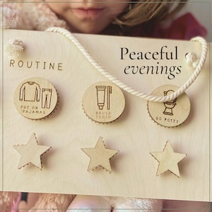 May include: A light-colored wooden routine board with the text Peaceful evenings and ROUTINE. It features three circular wooden tokens with icons for pajamas, brushing teeth, and using the potty, plus three wooden star shapes and a white rope.