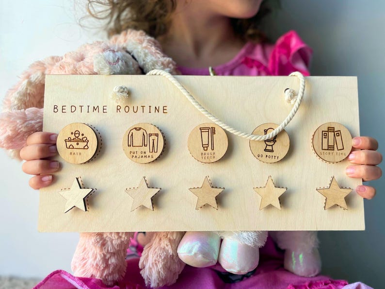 May include: Wooden bedtime routine chart with five circular icons for bath, pajamas, brushing teeth, potty, and story time. Includes five star-shaped reward tokens. The chart is held by a person.