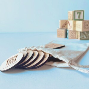 May include: A set of round, light-colored wooden discs with engraved icons and text, including one labeled Play-Doh, partially emerging from a cream-colored fabric drawstring pouch. In the background, a stack of wooden alphabet and number blocks.