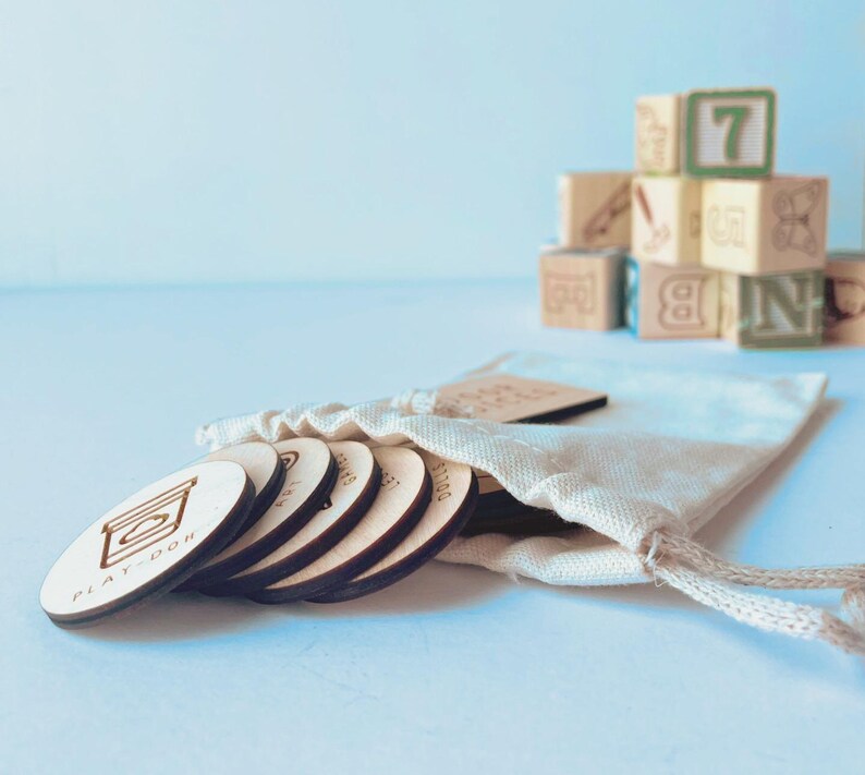 May include: A set of round, light-colored wooden discs with engraved icons and text, including one labeled Play-Doh, partially emerging from a cream-colored fabric drawstring pouch. In the background, a stack of wooden alphabet and number blocks.
