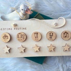 May include: Wooden bedtime routine chart with engraved icons for bath, pajamas, brushing teeth, potty, and story time. Includes star-shaped reward tokens. A decorative duck and a book are in the background.
