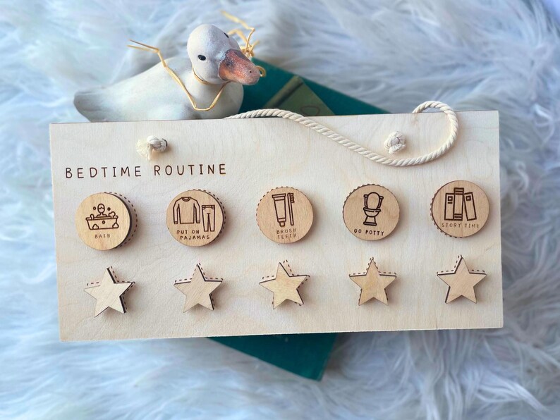 May include: Wooden bedtime routine chart with engraved icons for bath, pajamas, brushing teeth, potty, and story time. Includes star-shaped reward tokens. A decorative duck and a book are in the background.