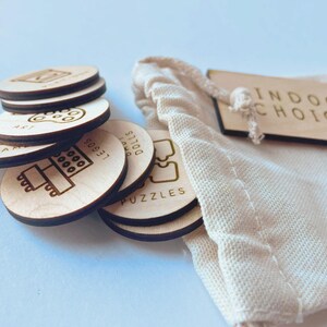 May include: A set of round light-colored wooden tokens with engraved icons and text including Art, Legos, Dolls, and Puzzles, stacked next to a cream-colored fabric drawstring bag with a wooden tag labeled Indoor Choices.