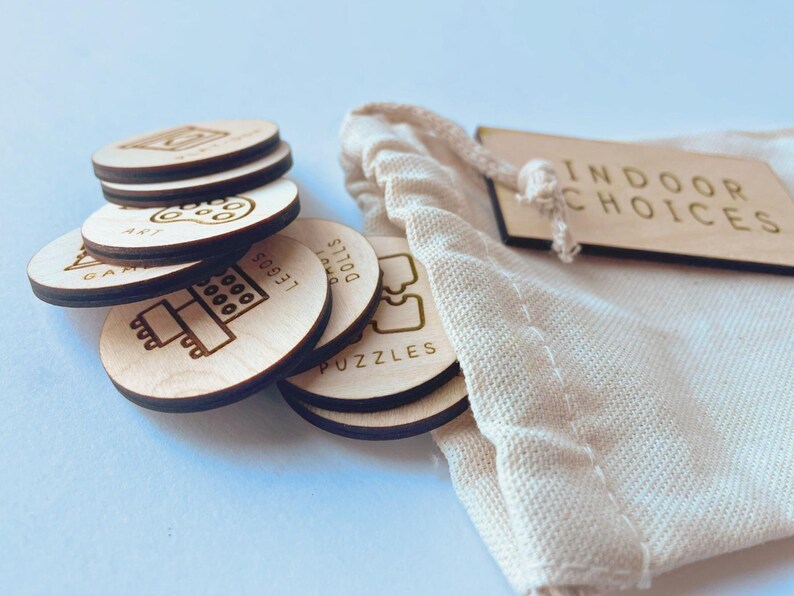 May include: A set of round light-colored wooden tokens with engraved icons and text including Art, Legos, Dolls, and Puzzles, stacked next to a cream-colored fabric drawstring bag with a wooden tag labeled Indoor Choices.