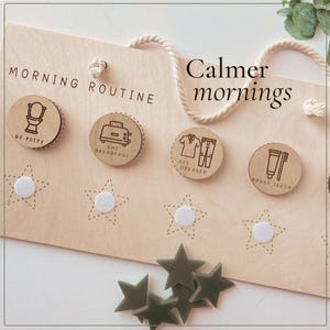 May include: A light-colored wooden morning routine board with a white rope hanger. It features four circular wooden tokens with icons and text: Go Potty, Eat Breakfast, Get Dressed, and Brush Teeth. Below are four white velcro stars and loose olive stars.