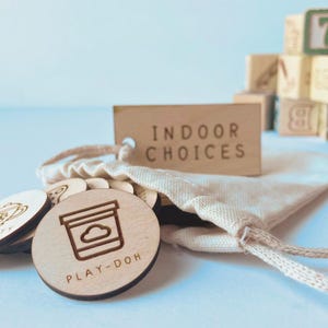 May include: A set of round wooden tokens with laser-engraved icons and text including "PLAY-DOH" and "TEA PARTY" spilling from a small beige cotton drawstring bag. A rectangular wooden tag labeled "INDOOR CHOICES" is attached to the bag.