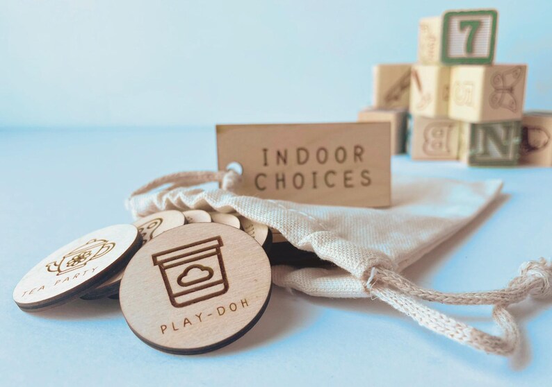 May include: A set of round wooden tokens with laser-engraved icons and text including "PLAY-DOH" and "TEA PARTY" spilling from a small beige cotton drawstring bag. A rectangular wooden tag labeled "INDOOR CHOICES" is attached to the bag.