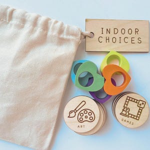 May include: A beige cotton drawstring bag, a rectangular wooden tag labeled INDOOR CHOICES, several colorful heart-shaped rings in blue, green, orange, and purple, and round wooden tokens featuring engraved icons for ART and GAMES.