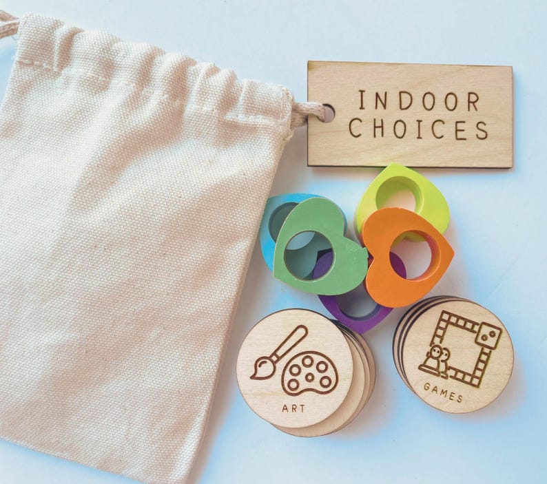 May include: A beige cotton drawstring bag, a rectangular wooden tag labeled INDOOR CHOICES, several colorful heart-shaped rings in blue, green, orange, and purple, and round wooden tokens featuring engraved icons for ART and GAMES.