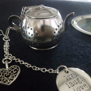 May include: A stainless steel tea infuser in the shape of a teapot, with a small matching tray. The infuser has a chain with a heart-shaped charm and a round charm that says "live laugh love". The teapot infuser is approximately 7.6 cm tall.