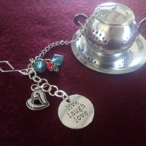 Könnte beinhalten: Silberfarbener Tee-Infuser in Teekannenform, auf einer passenden Untertasse. Eine Kette verbindet den Infuser mit einem Anhänger mit den Worten "live laugh love" und einem Herz-Anhänger. Dekorative türkisfarbene und rote Perlen.