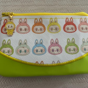 May include: A rectangular cosmetic bag with a white background featuring repeating cartoon bunny faces in various colors. The bottom is lime green, and the zipper is gold with a charm. The bag is approximately 8 inches wide.