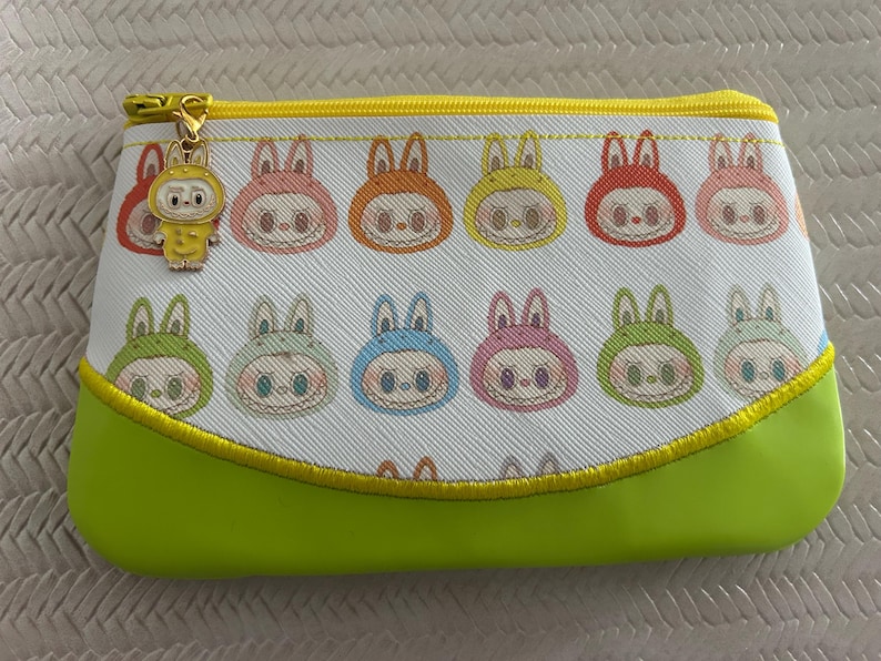 May include: A rectangular cosmetic bag with a white background featuring repeating cartoon bunny faces in various colors. The bottom is lime green, and the zipper is gold with a charm. The bag is approximately 8 inches wide.