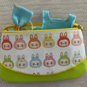 May include: A rectangular, zippered pouch with a white background and a repeating pattern of cartoon bunny faces in various colors. The pouch has a lime green base and a yellow zipper. A light blue bow and a small blue garment are visible inside.