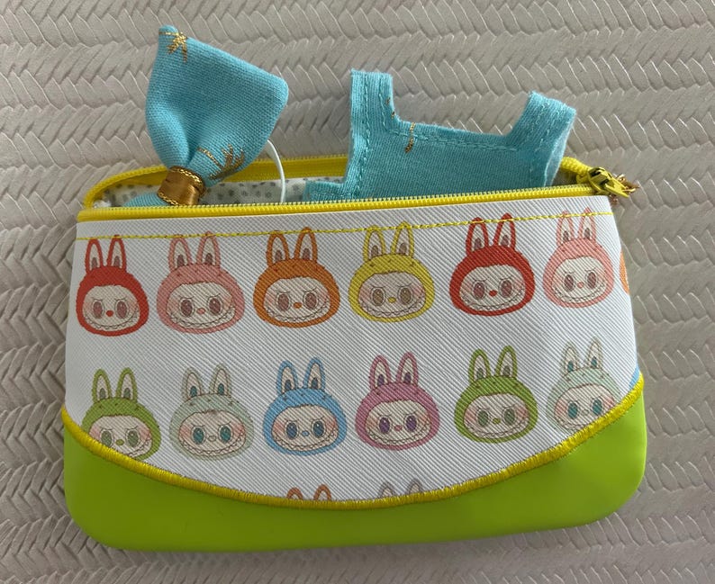 May include: A rectangular, zippered pouch with a white background and a repeating pattern of cartoon bunny faces in various colors. The pouch has a lime green base and a yellow zipper. A light blue bow and a small blue garment are visible inside.