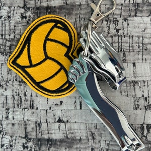 May include: A yellow heart-shaped keychain with a black outline of a volleyball. The keychain is attached to a silver nail clipper with a small silver loop.
