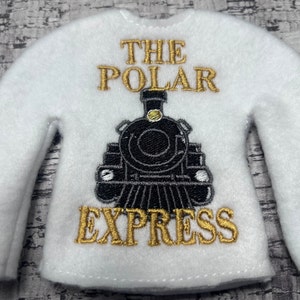 May include: A white long-sleeved shirt with the words "THE POLAR EXPRESS" embroidered in gold thread. A black train is embroidered below the words. The shirt is designed for a small doll or figure.
