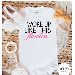 May include: White baby onesie with the text "I WOKE UP LIKE THIS flawless" in black and pink. The onesie is surrounded by baby accessories, including white moccasins, a wooden teether, and a mustard-colored bow.