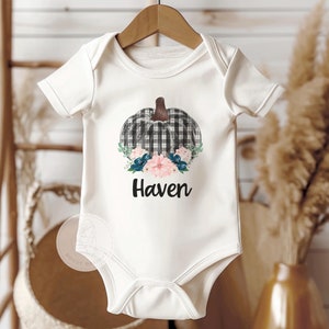 Könnte beinhalten: Weißer Baby-Strampler mit einem schwarz-weiß karierten Kürbis mit blauen und rosa Blumen und dem Namen "Haven" darunter.