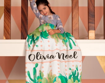 May include: A white and pink blanket with a green cactus pattern and white polka dots. The blanket has the name "Olivia Noel" written in black cursive on a pink stripe.