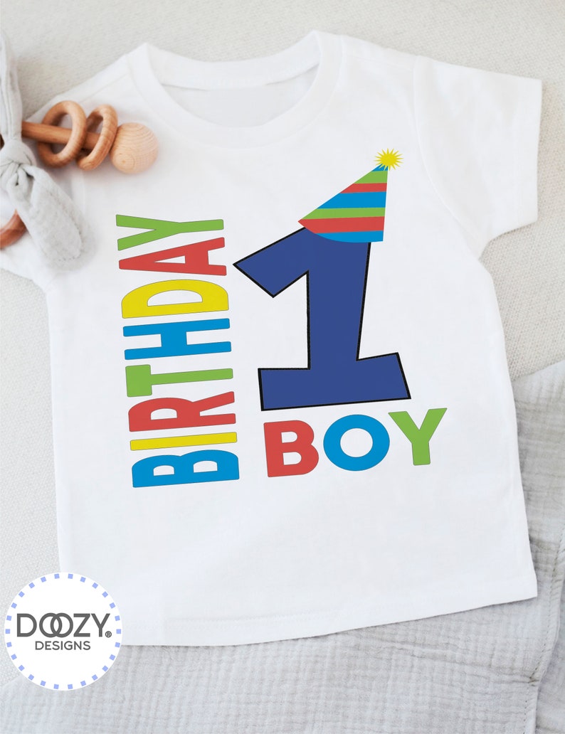 May include: White short-sleeved t-shirt with a blue number one wearing a multi-colored party hat. The text "Birthday Boy" is written in colorful letters.
