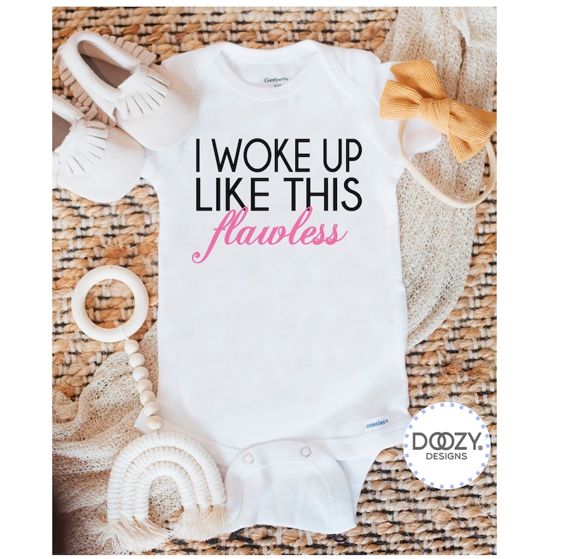 May include: White baby onesie with the text "I WOKE UP LIKE THIS flawless" in black and pink. The onesie is surrounded by baby accessories, including white moccasins, a wooden teether, and a mustard-colored bow.