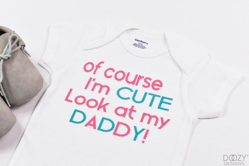 May include: White baby onesie with the text "of course I'm CUTE look at my DADDY!" in pink and teal.