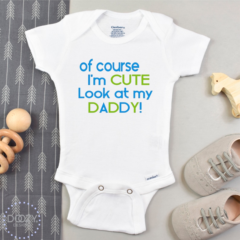 May include: White baby onesie with blue and green text that reads "of course I'm CUTE Look at my DADDY!"