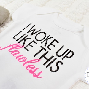 May include: White baby onesie with the text "I WOKE UP LIKE THIS Flawless" in black and pink. The onesie is on a white, textured surface. A pair of gray baby shoes and a wooden toy are in the background.