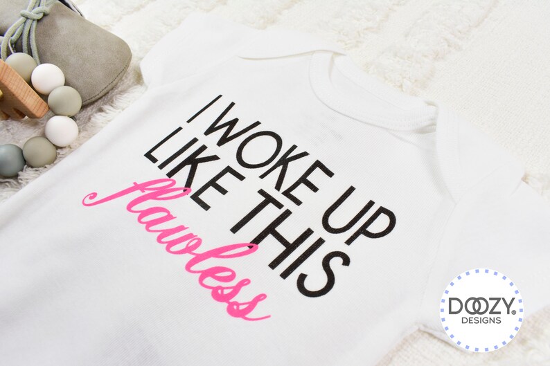 May include: White baby onesie with the text "I WOKE UP LIKE THIS Flawless" in black and pink. The onesie is on a white, textured surface. A pair of gray baby shoes and a wooden toy are in the background.