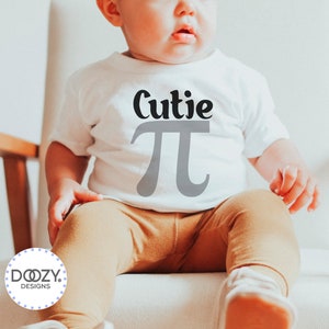 Pu&ograve; includere: Un body bianco per neonato con il testo "Cutie Pi" in lettere nere e grigie. Il body &egrave; indossato da un bambino seduto su una sedia bianca.