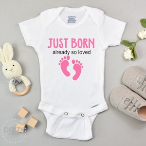 May include: White baby onesie with short sleeves and pink footprints. The onesie has the text "JUST BORN already so loved" printed on it.