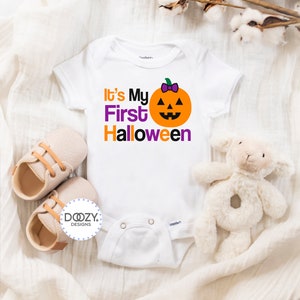Puede incluir: Un body blanco para beb&eacute; con el texto "It's My First Halloween" en letras naranja y morado. Una peque&ntilde;a calabaza naranja con un lazo morado y una cara sonriente est&aacute; encima del texto.