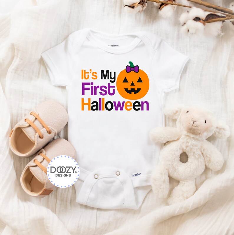 Puede incluir: Un body blanco para beb&eacute; con el texto "It's My First Halloween" en letras naranja y morado. Una peque&ntilde;a calabaza naranja con un lazo morado y una cara sonriente est&aacute; encima del texto.