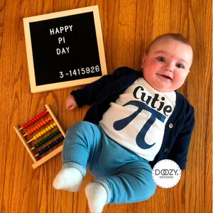 Pu&ograve; includere: Un bambino che indossa un body bianco con una grafica blu "Cutie Pi" e jeans blu. Il bambino &egrave; sdraiato su un pavimento di legno con un pannello di lettere che dice "Happy Pi Day 3-1415926" e un abaco di legno con perline colorate.